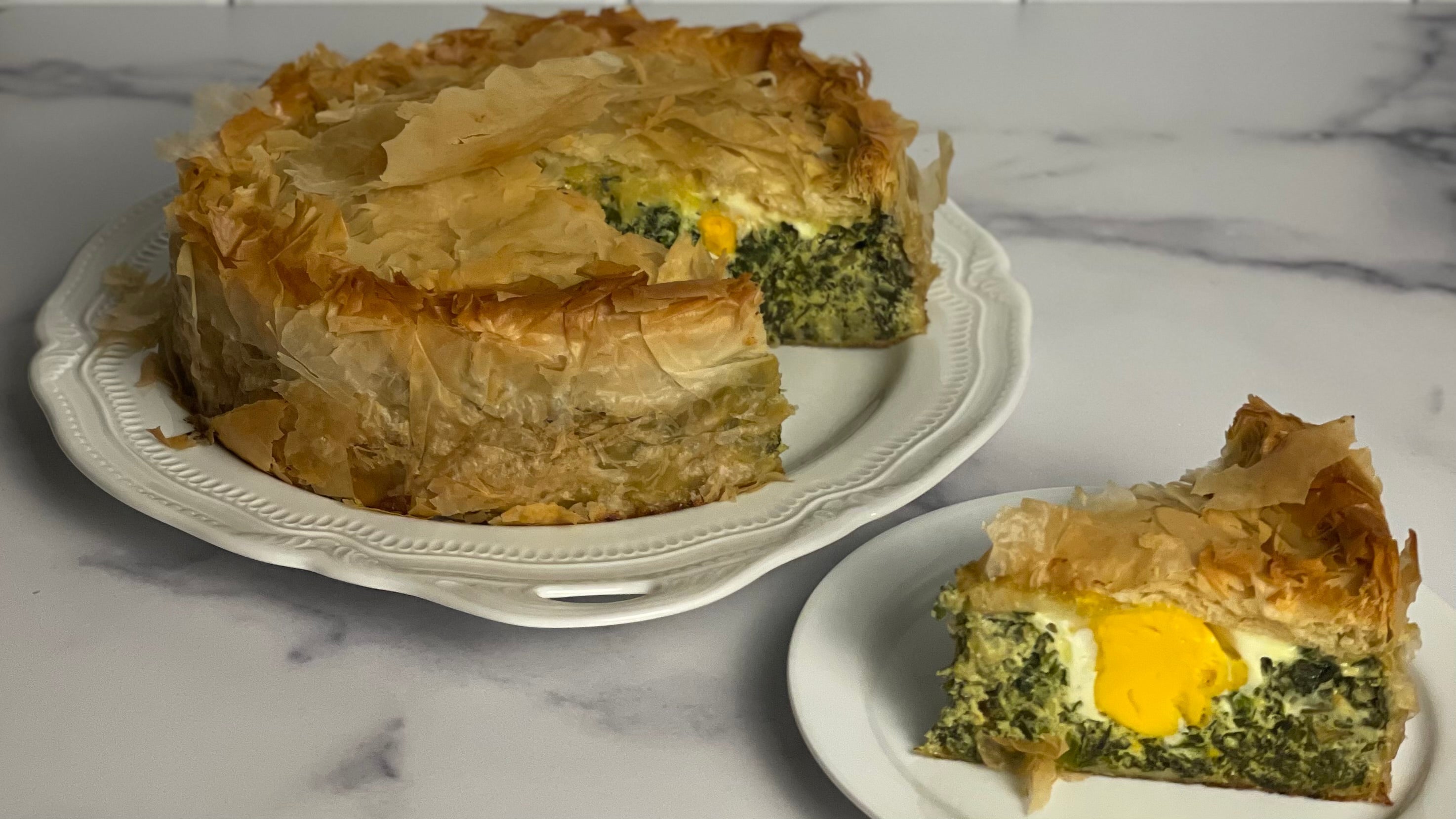 A festive phyllo covered savory "cake" shows one slice revealing cooked leafy greens and a whole egg.