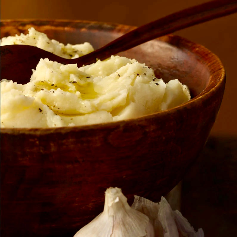 A dark wooden bowl is filled with fluffy mashed potatoes. You can see olive oil and pepper on top.