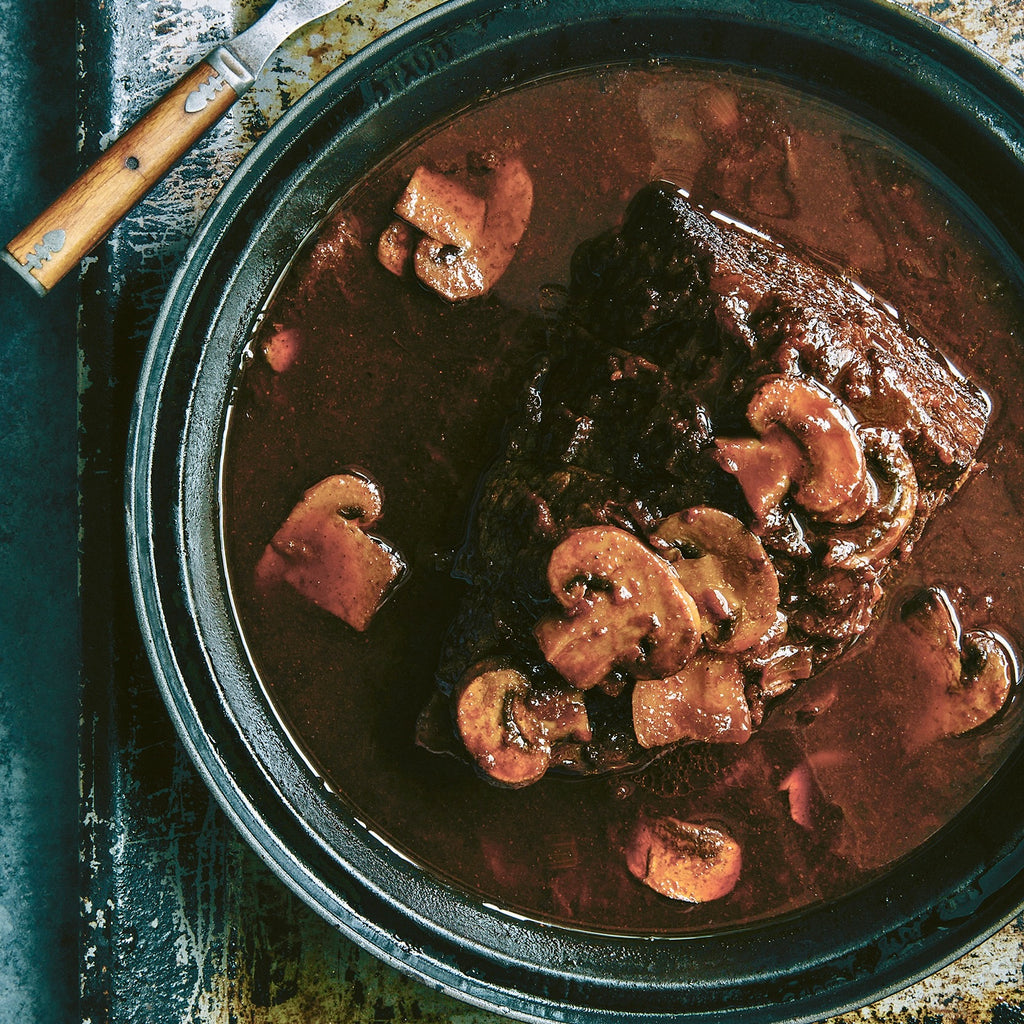 4 lbs of roasted beef sits in round dutch oven covered in a rich broth with sliced mushroom.