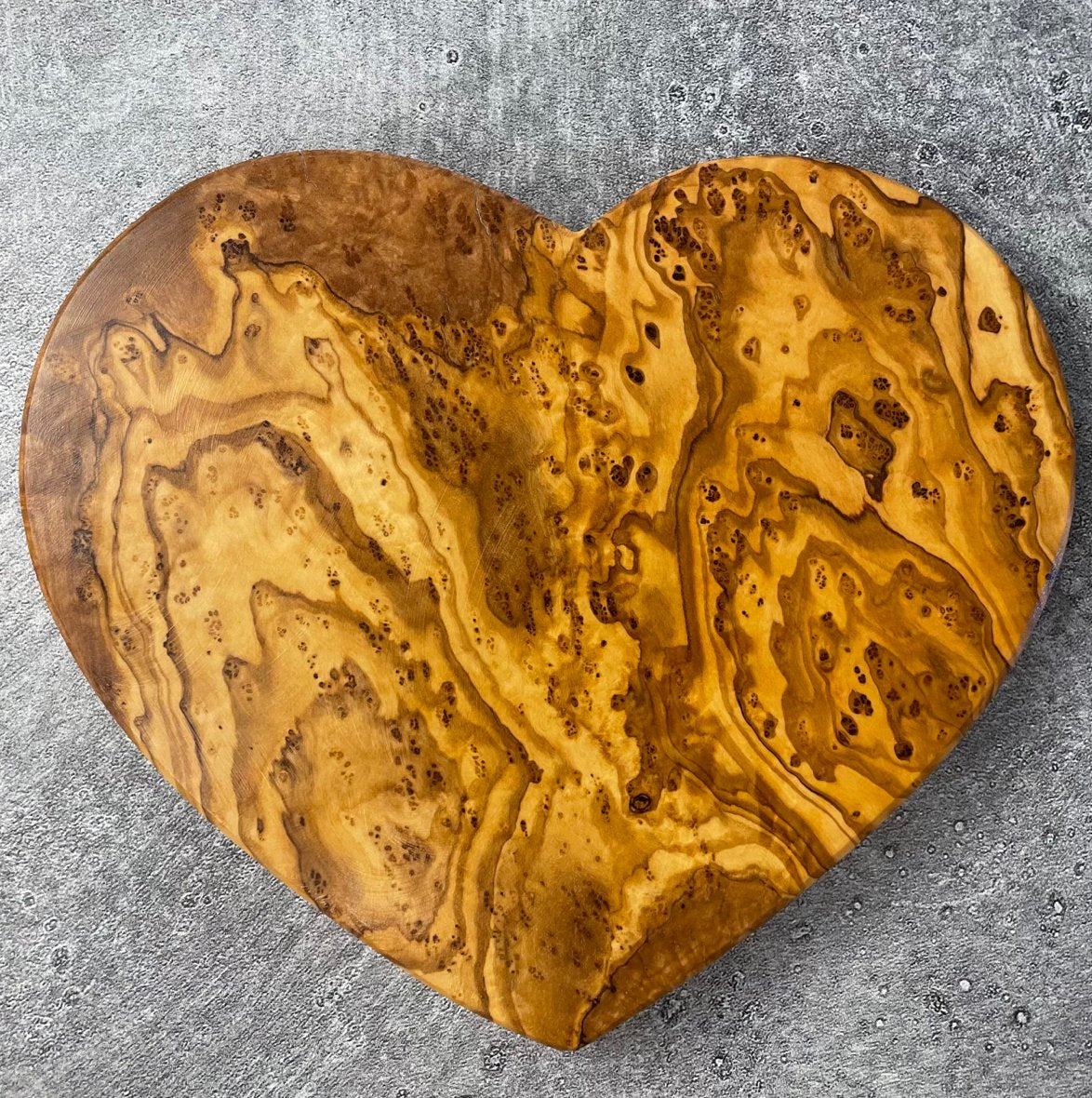 Heart-shaped olivewood board is shown on a stone countertop. The wood grain is elaborating and beautifully figured.