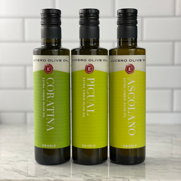 Three single variety olive oils are shown on a white marble table with white tile background. Left to Right:  Coratina, Picual, and Ascolano