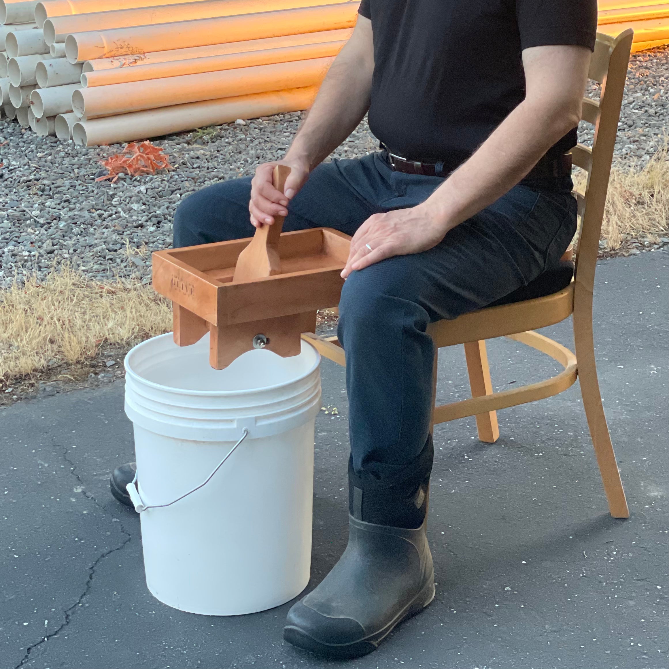 Man sits on the American Olive Farmer wooden olive crusher which is placed on a kitchen chair. There is a 5 gallon bucket below the device which shows how olives drop into the bucket below.