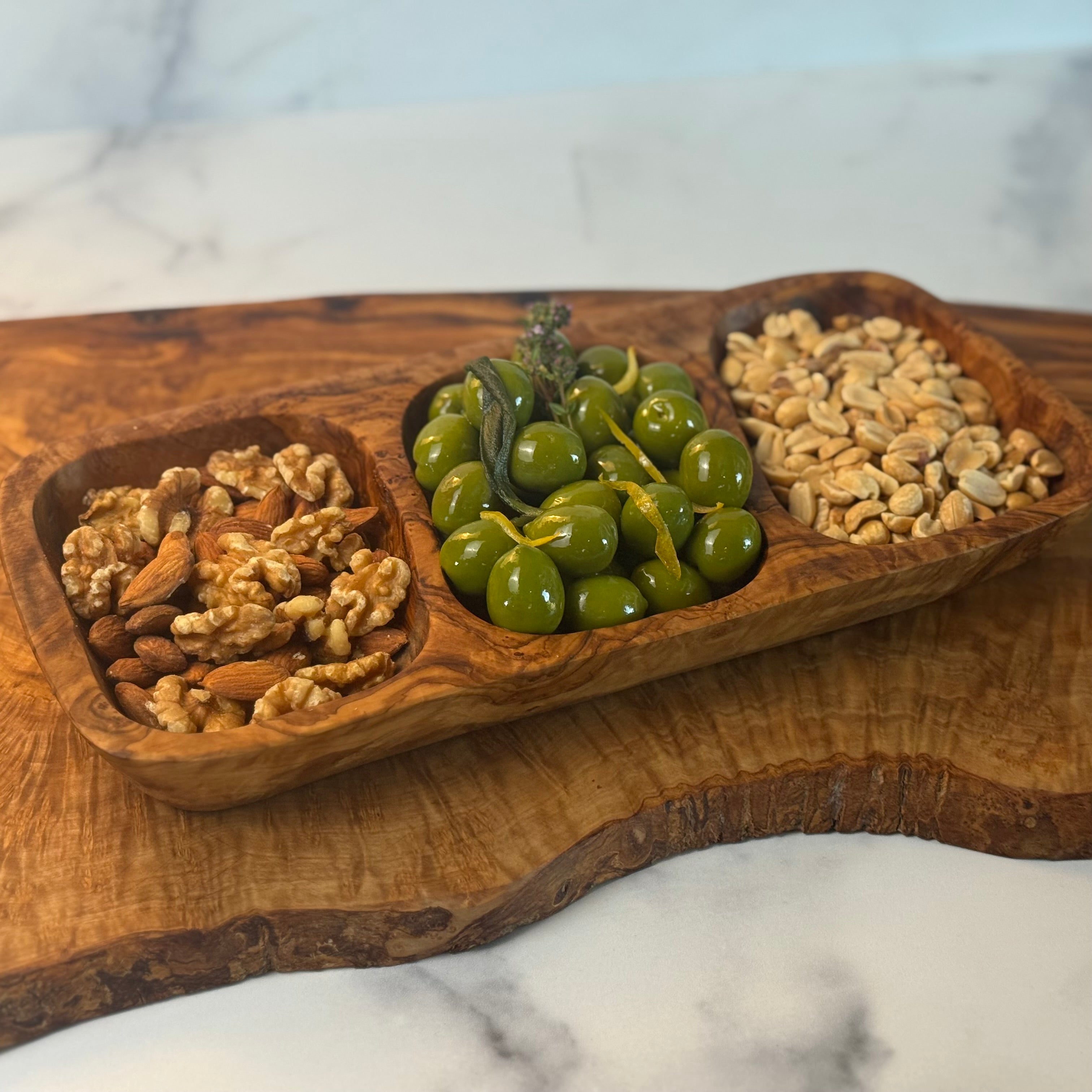 An olivewood appetizer tray is shown sitting on another piece of olivewood and both are on a marble counter. There are three sections hollowed out on the tray and filled with (left to right) a walnut & almond mix, herbed olives, and roasted peanuts.