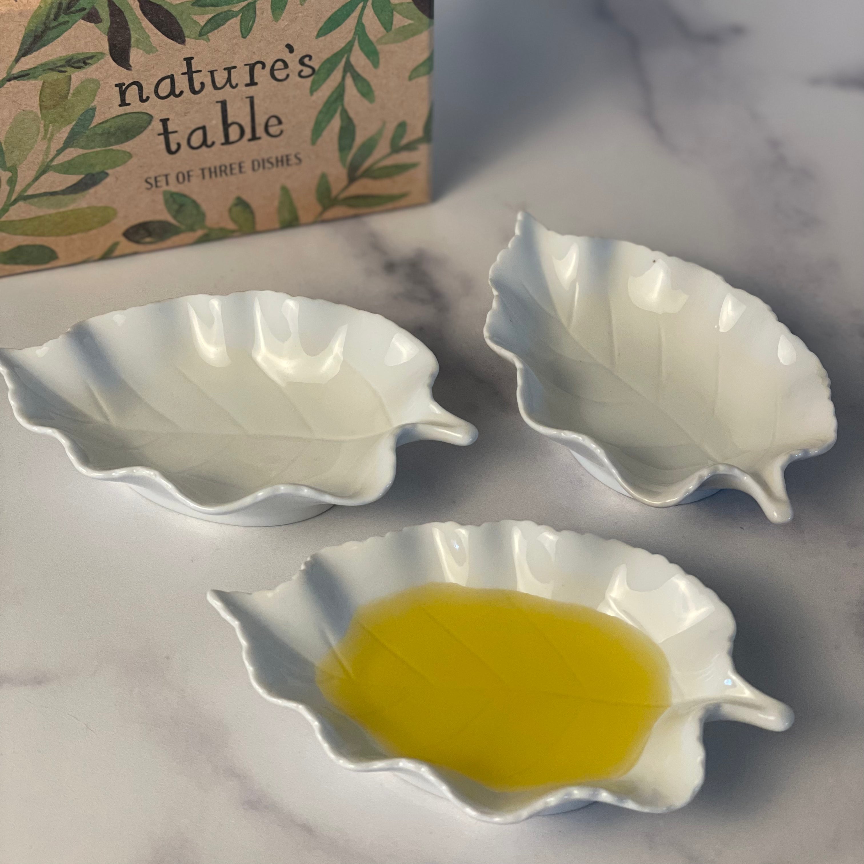 3 delicate porcelain dipping dishes are shown close up on a marble counter. One is filled with Lucero Olive Oil. A decorative gift box is in the background.