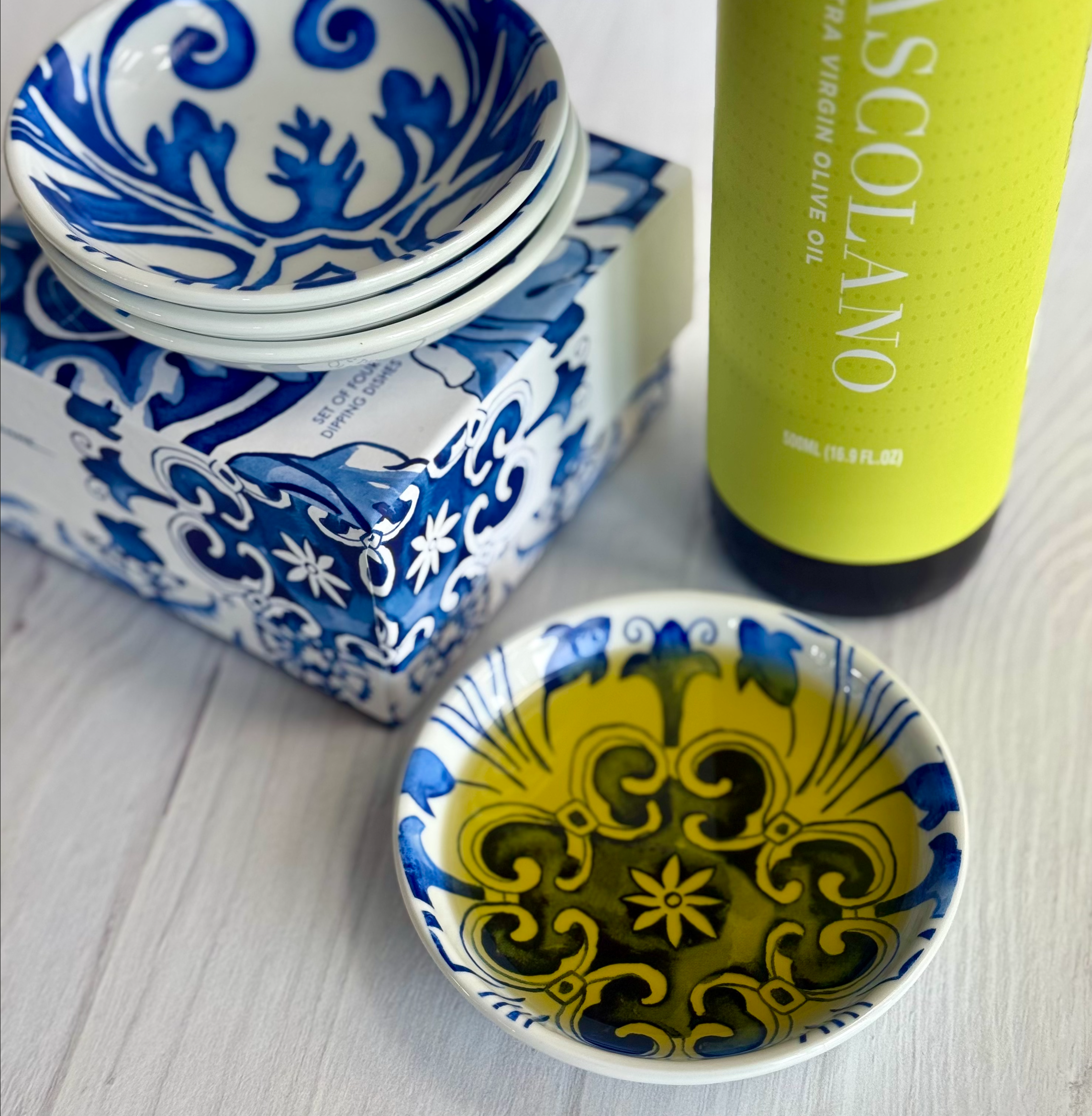 Four assorted dipping dishes with stunning Mediterranean tile work decor are shown with a decorative gift box. One of the dishes is filled with oil, and a bottle of Lucero Ascolano is shown just out of frame.