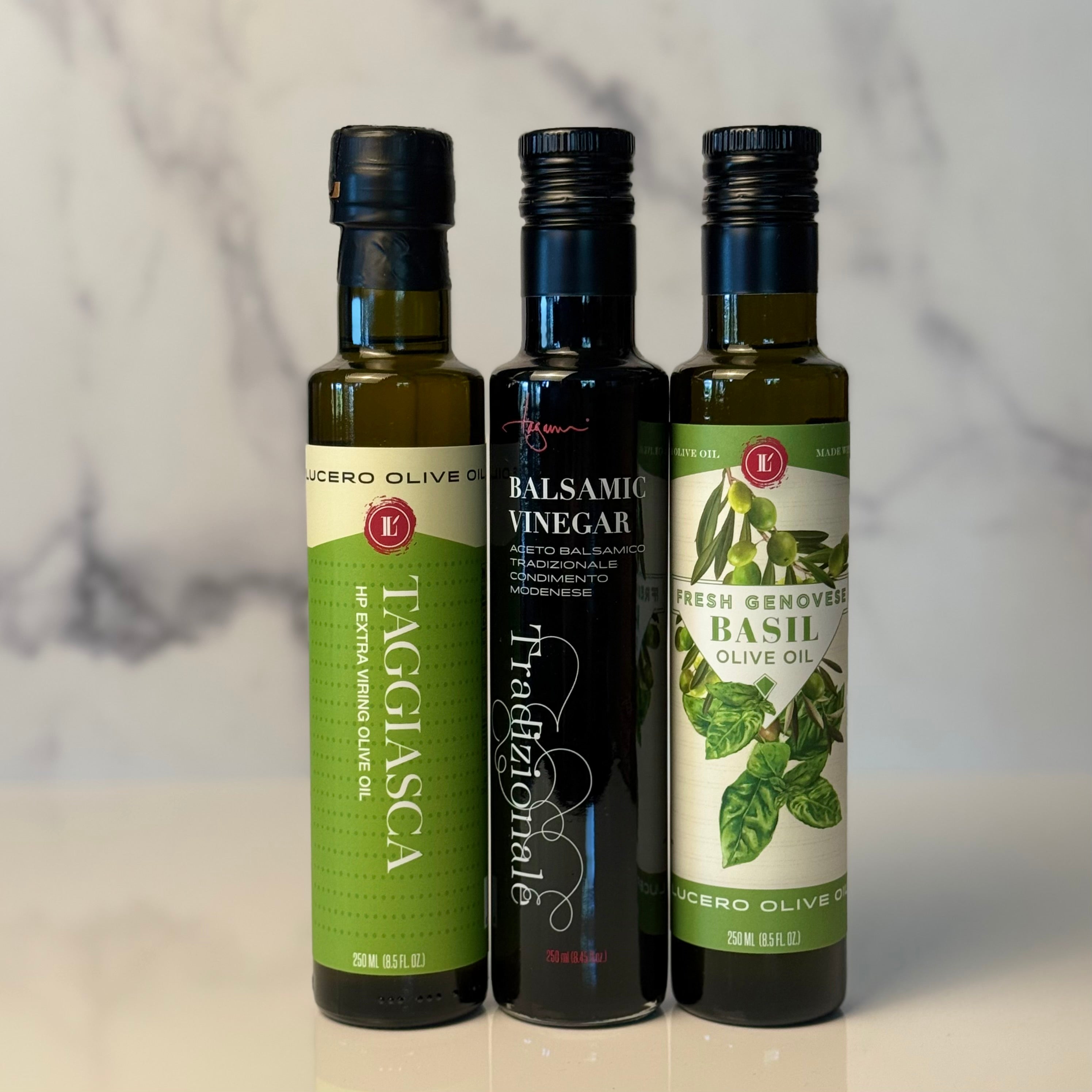 Three small bottles of olive oil and vinegar from Lucero Olive Oil stand on a white marble surface against a white marble background. The bottles are labeled Taggasca, Balsamic Vinegar, and Fresh Genovese Basil Olive Oil.