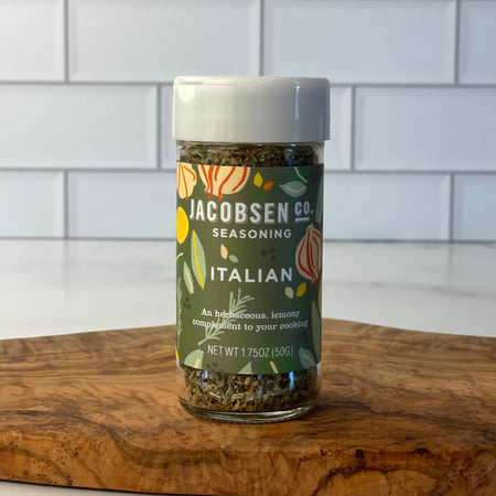 Close up image of a jar of Jacobsen Co. Italian seasoning on an olivewood board in a marble and tile kitchen