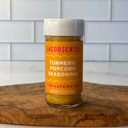 Single jar of Turmeric Popcorn seasoning is on an Olivewood board in a white tile kitchen