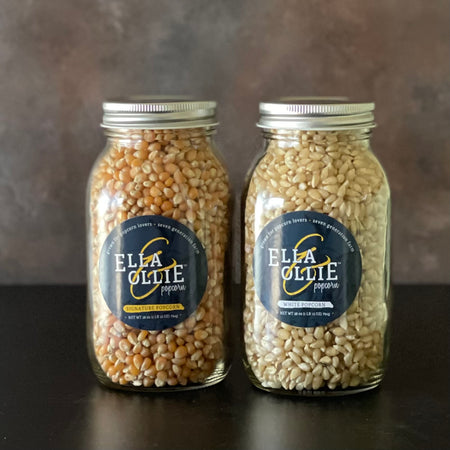 2 mason jars are shown on a simple counter, one is filled with Ella & Ollie Signature popcorn, the other with the company's white popcorn