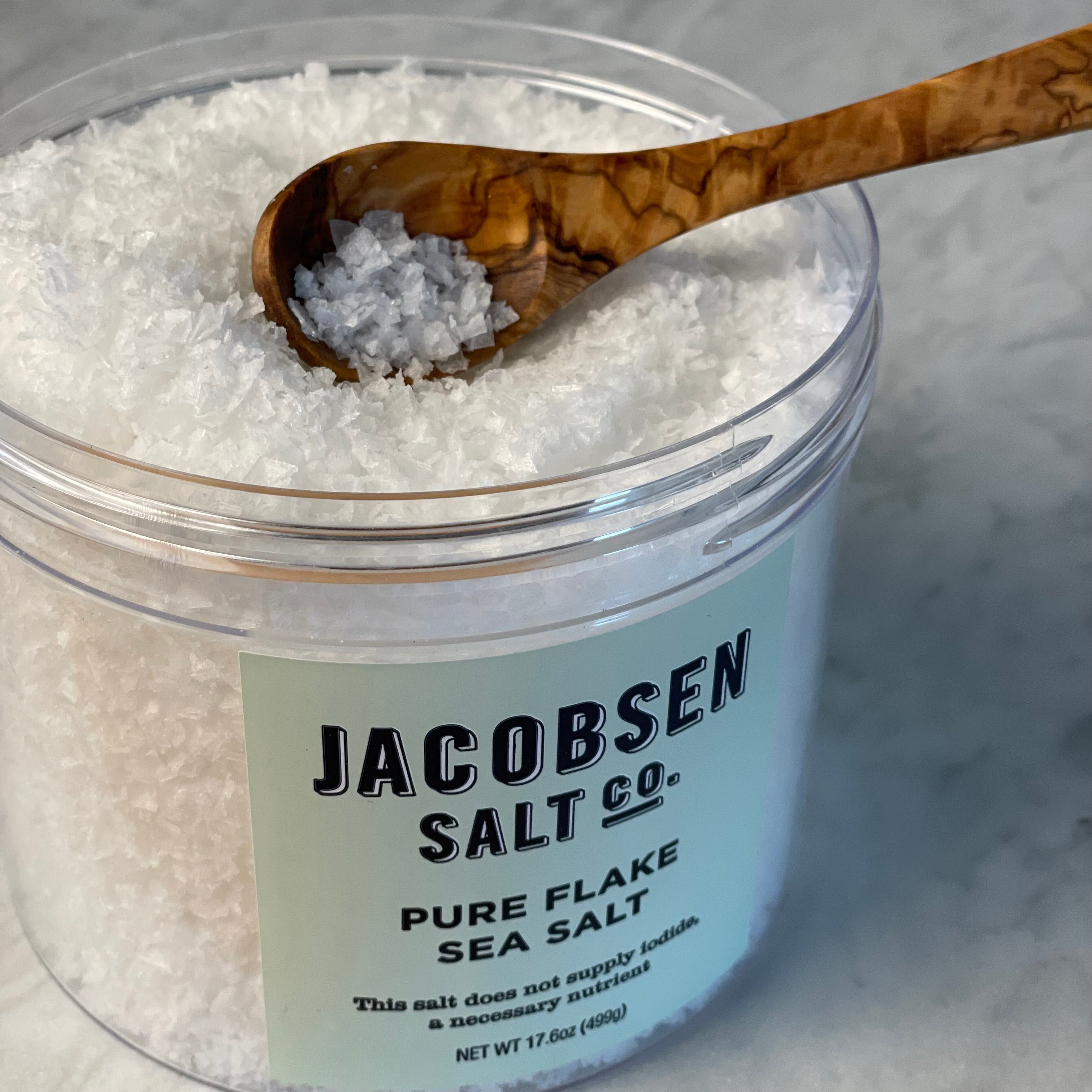 Close up of a 499 gm  17.6 oz jar of pure flake salt from Jacobsen Salt Co. shown with the lid off. A small olivewood condiment spoon scoops a few flakes.