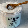 Close up of a 499 gm  17.6 oz jar of pure flake salt from Jacobsen Salt Co. shown with the lid off. A small olivewood condiment spoon scoops a few flakes.