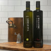 Tall bottle of Arbequina EVOO and its matching bottle of Balsamic Vinegar stand side by side in a tile kitchen on an olivewood cutting board.