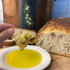 Liz's hand is shown holding a chuck of freshly baked bread drenched with Olio Novello dripping onto a porcelain plate. A cut loaf and bottle of Olio Novello are in the background.