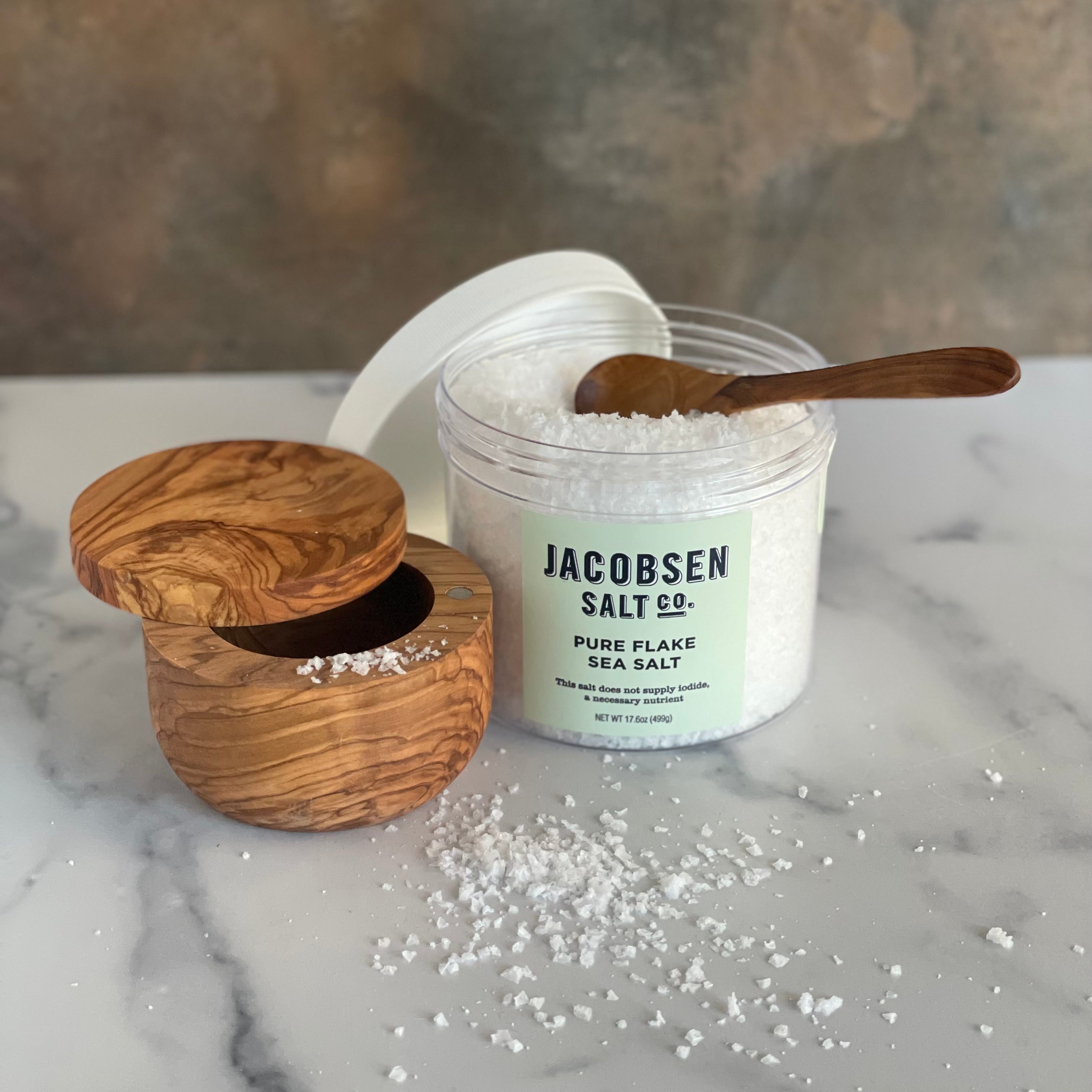 An olivewood salt cellar, 499 gm jar of flake salt, and olivewood condiment spoon are shown on a marble board.