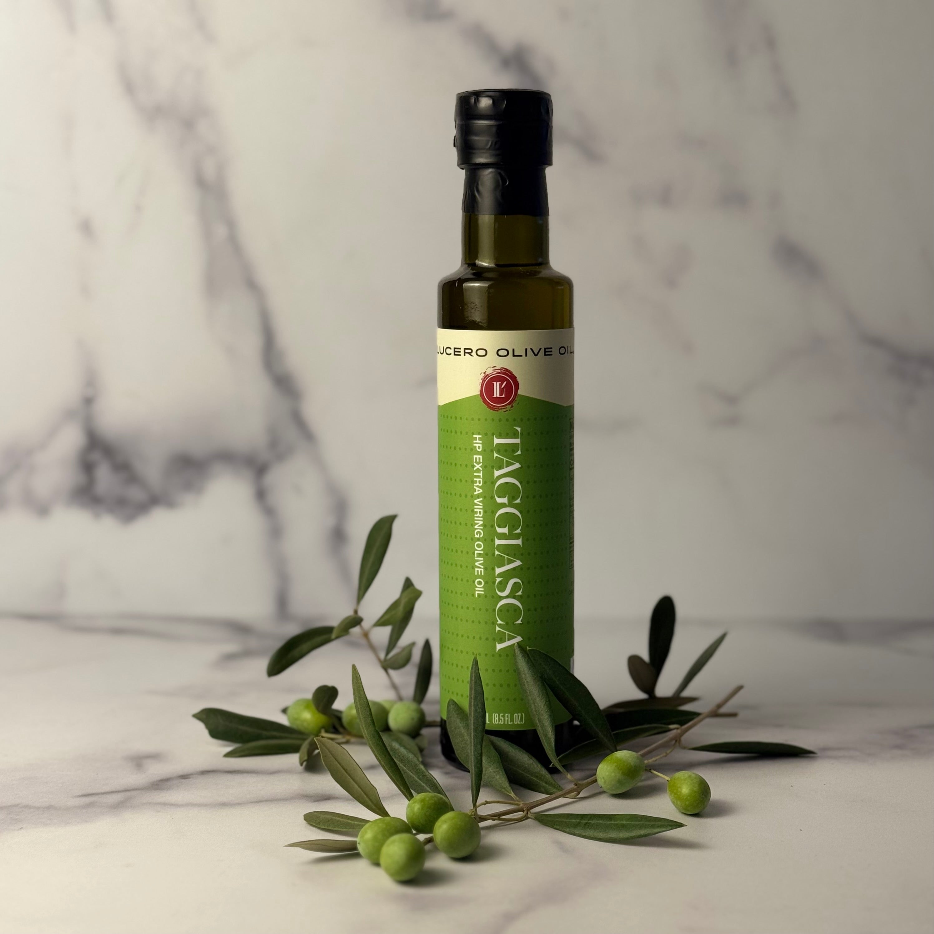 Single bottle of Lucero Taggiasca Olive OIl surrounded by fresh green Taggiasca olives