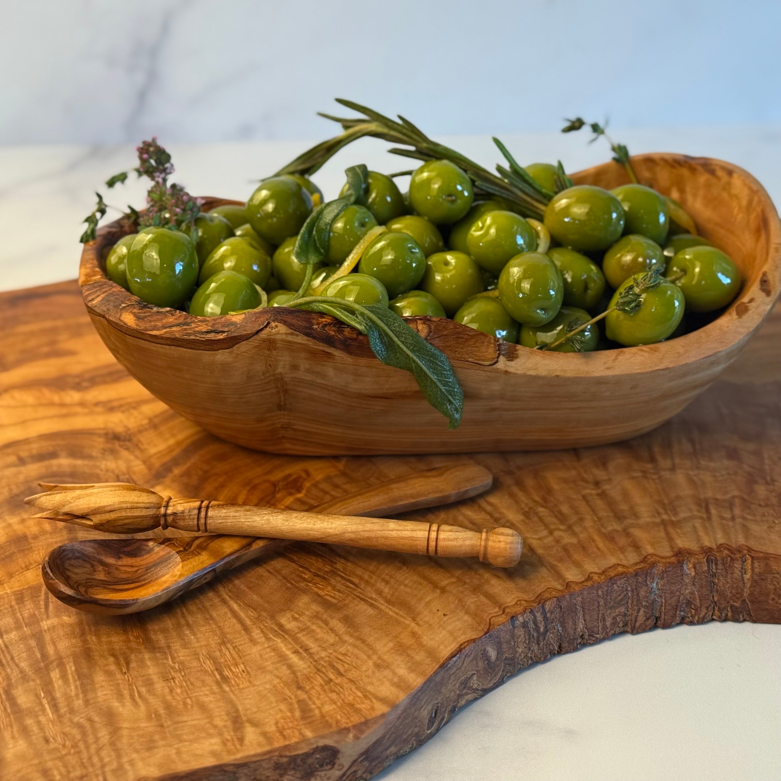 Wooden bowl filled with green olives on a wooden surface with two mini serving utensils, a condiment spoon and a classic olive pick.