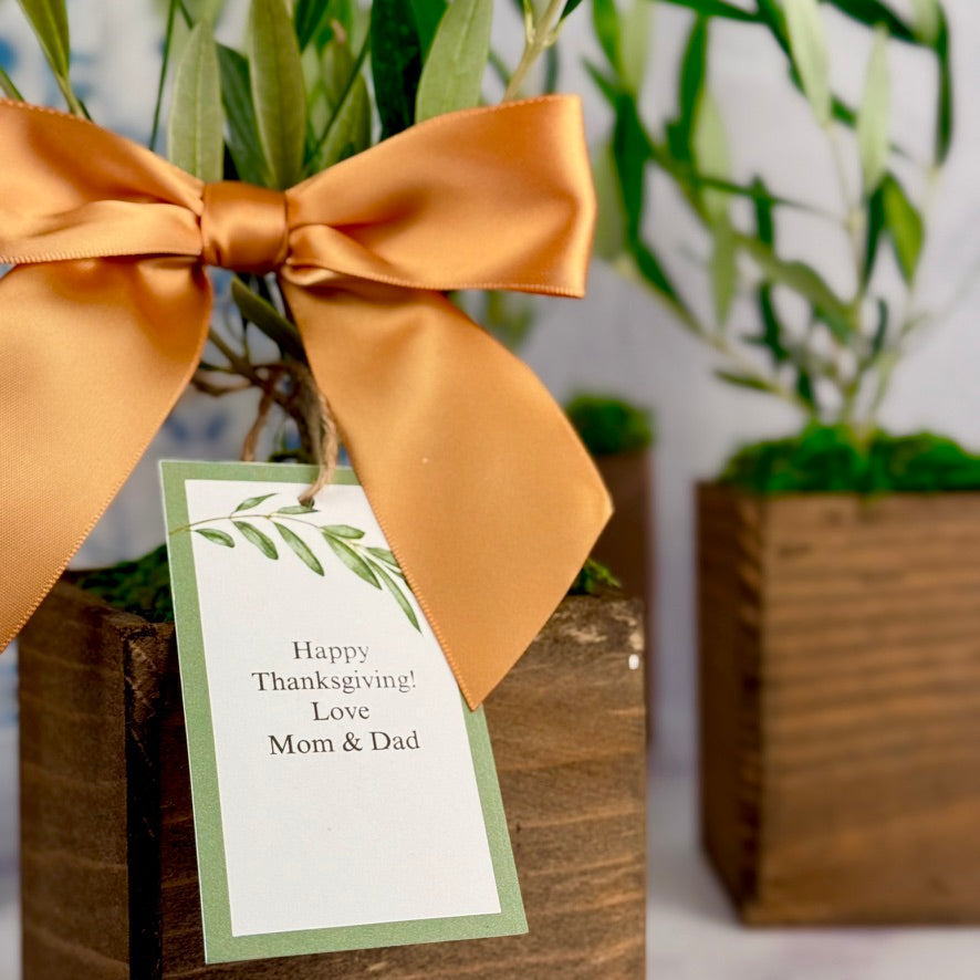 This is a close up view of a gift tag, which says, "Happy Thanksgiving! Love Mom & Dad" with is tied to the tree with jute string. There is a satin bow and the entire tree is in a rustic wooden square cachepot.