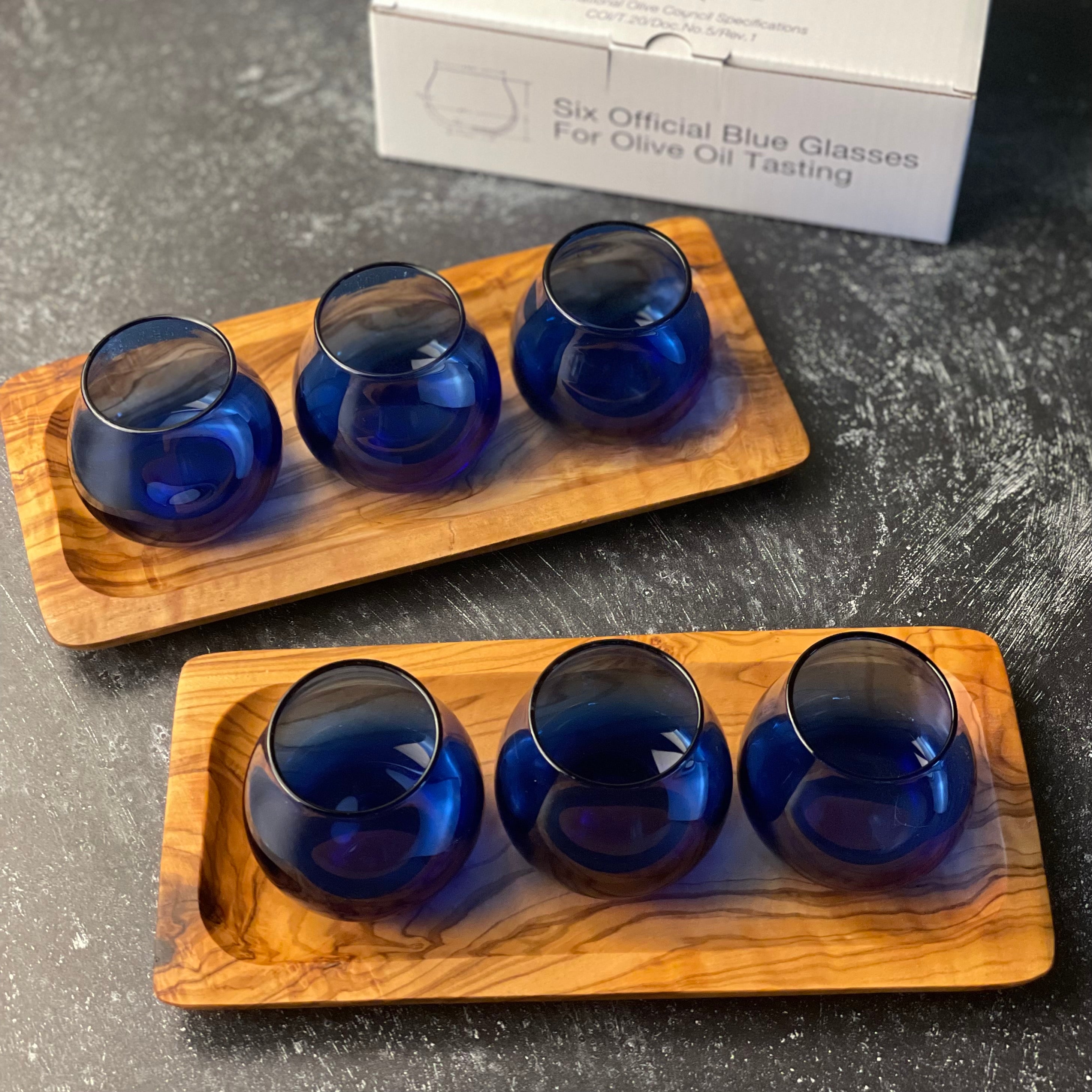 A set of six olive oil tasting glasses are shown on two olive wood trays, three glasses on each tray.  