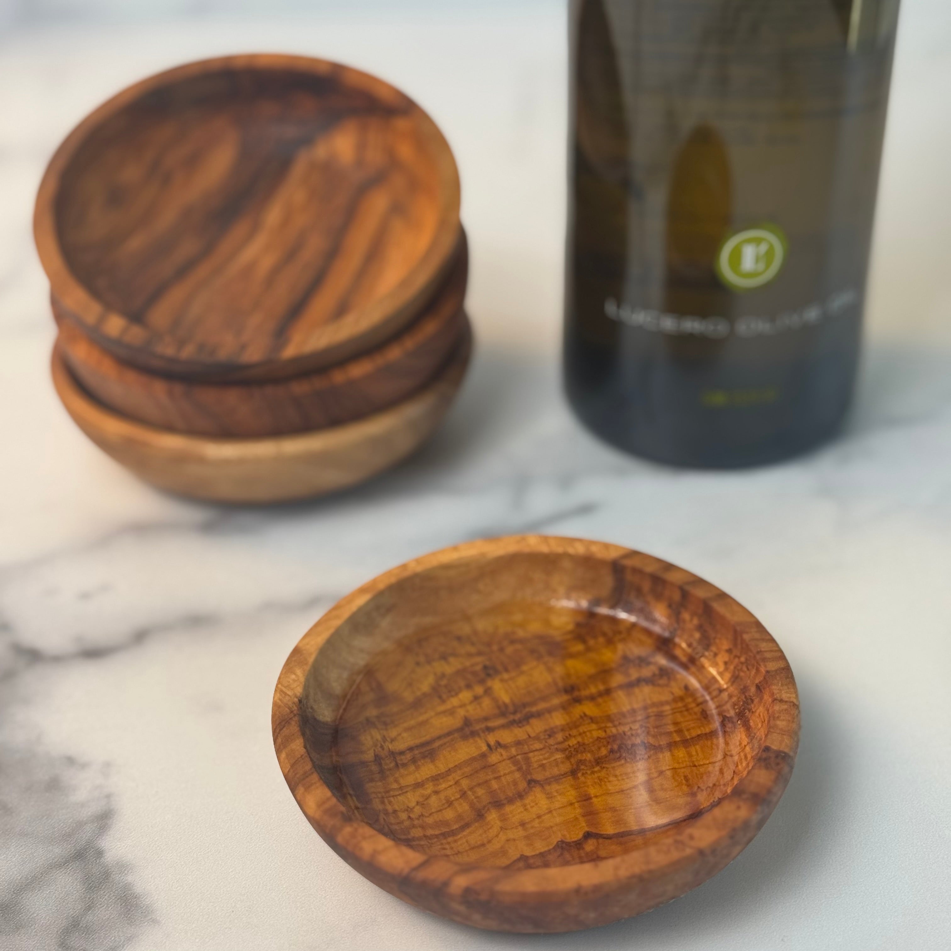 Three shallow olivewood dipping bowls are stacked next to a bottle of Arbequina and a fourth bowl rests in front filled with olive oil.