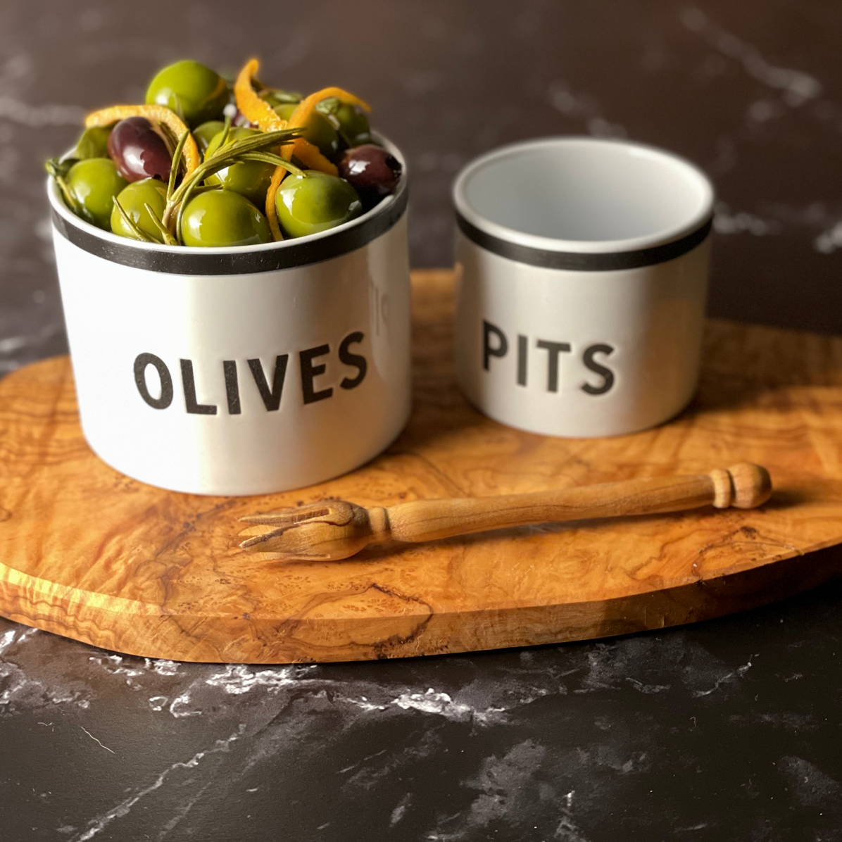 A small olivewood board, set of modern ceramic bowls, and dainty olive pick are shown on a black marble surface. The larger of the two bowls is filled with two types of olives.