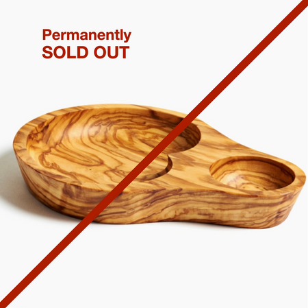 Image of a small, shallow plate meant to hold a small jar of olives. It is made of olivewood and here is a separate carve out for olive pits. There is a large line drawn through ti and the words "Permanently Sold Out".