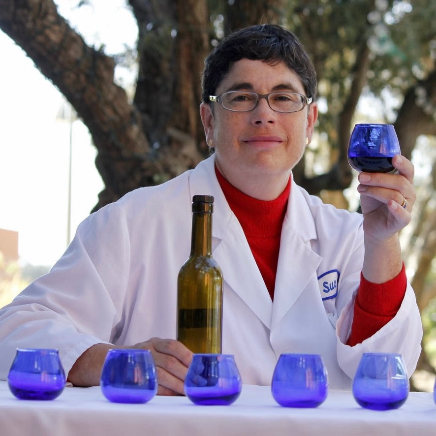 Woman in white lab coat raise blue tasting glass with five more glasses in front of her 