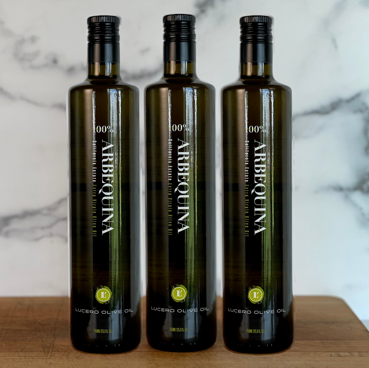 Three tall bottles of Lucero Arbequina Extra Virgin Olive Oil are arrayed on a wooden board in a marble kitchen