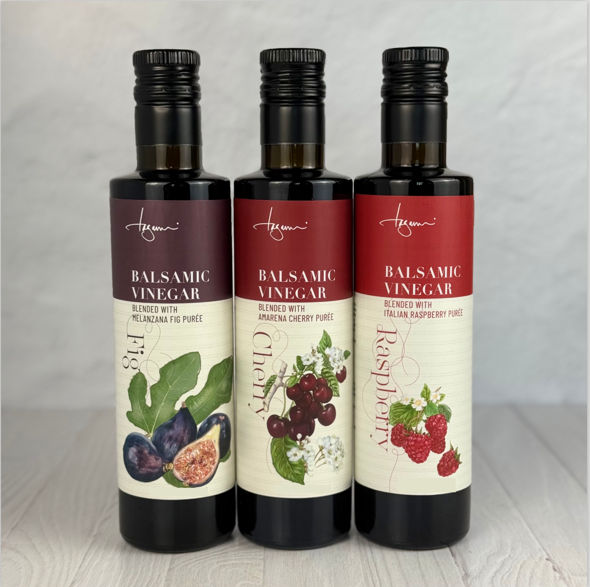 The new 250 ml bottles of Flavored balsamic Vinegar are shown lined up on a plain kitchen counter. Left to right: Melanzana Fig, Amarena Cherry, Italian Raspberry.