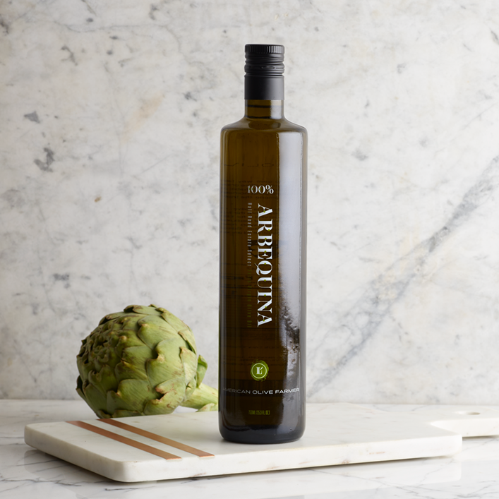 Tall unopened bottle of Arbequina Extra Virgin Olive Oil shown in a marble kitchen.