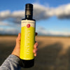 Liz holds a bottle of Ascolano at arm's length to capture its design with the golden light of dusk in front of a dry-farmed pasture.