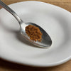 A silver spoon on a white plate holds the poultry and pork seasoning, which has a sandy texture.