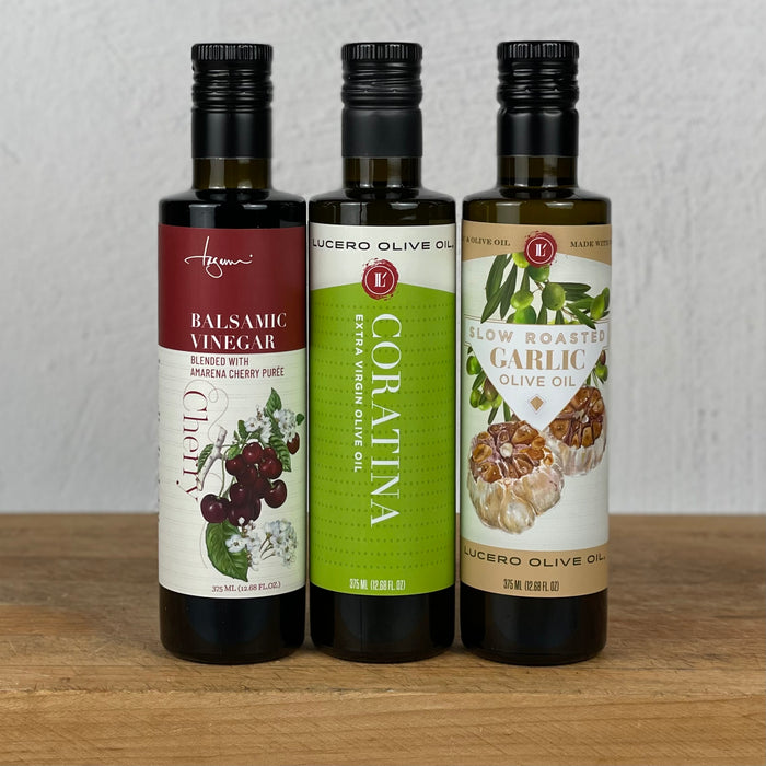 Three bottles are lined up left to right: Cherry Balsamic Vinegar, Coratina EVOO, and Roasted Garlic Olive Oil
