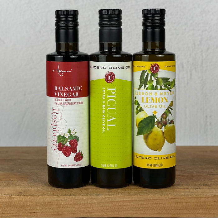 Three bottles are lined up left to right Raspberry Balsamic Vinegar, Picual EVOO, and Agrumato Lemon Olive Oil