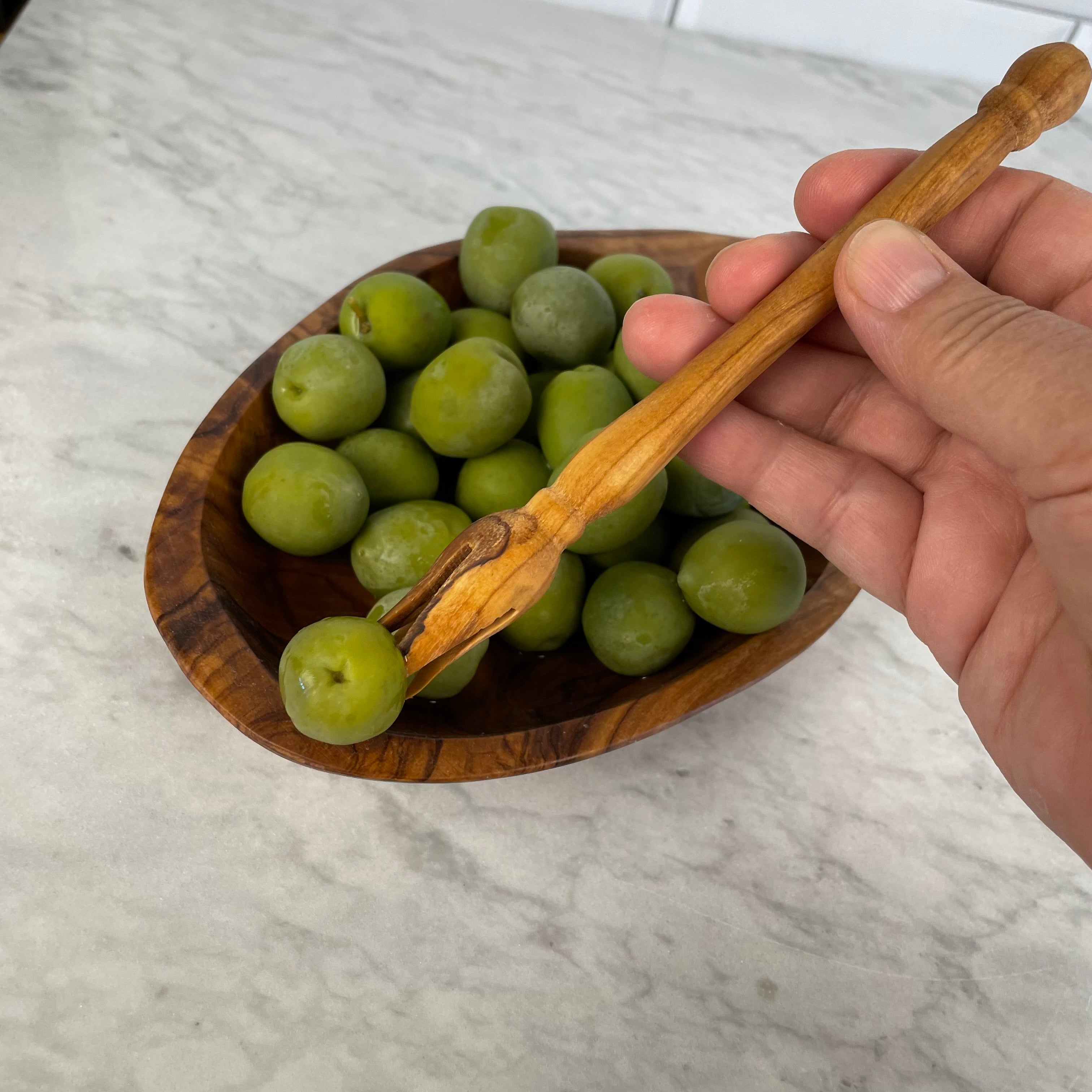 Close up of Liz's hand holding a single green olive with the pique (pick) over a small bowl olives to demonstrate use.