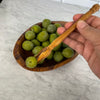 Close up of Liz's hand holding a single green olive with the pique (pick) over a small bowl olives to demonstrate use.