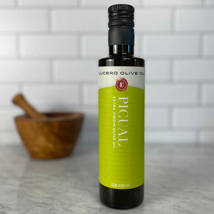 An unopened bottle of Picual EVOO on a white marble counter in front of white tile.