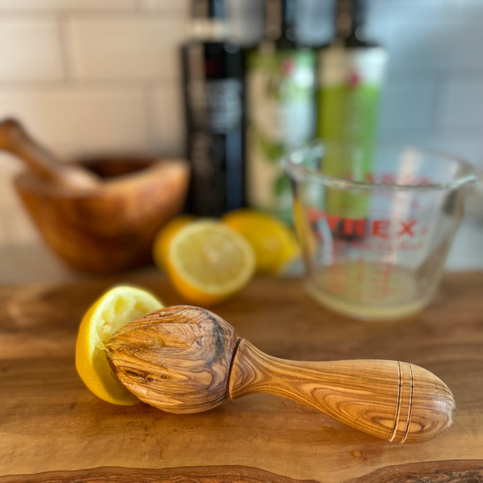 Handsome carved olivewood lemon reamer shown on a kitchen counter with reamed lemon half, cut lemons, and a glass measuring cup with lemon juice in the background