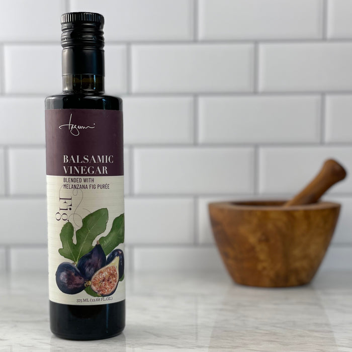 A bottle of Melanzana Fig Balsamic Vinegar is on a white marble counter in front of a white tile background.