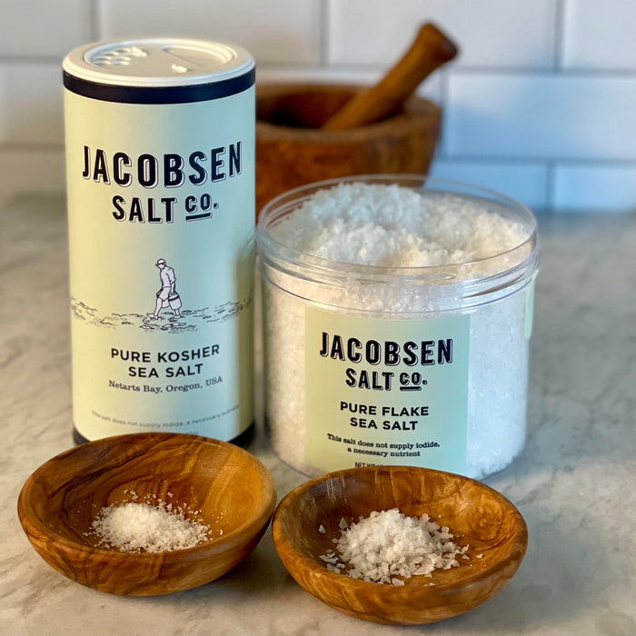 A canister of kosher salt is shown next to an open jar of flake salt. Small olivewood dishes in front show the difference in crystal size side-by-side.