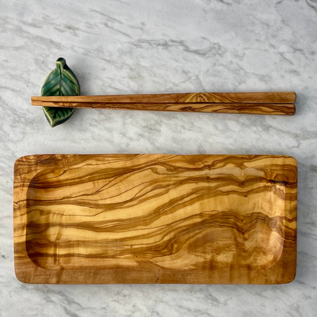 Rectangular olivewood service plate shown next to a pair of olivewood chopsticks which are resting on a green leaf chopstick holder