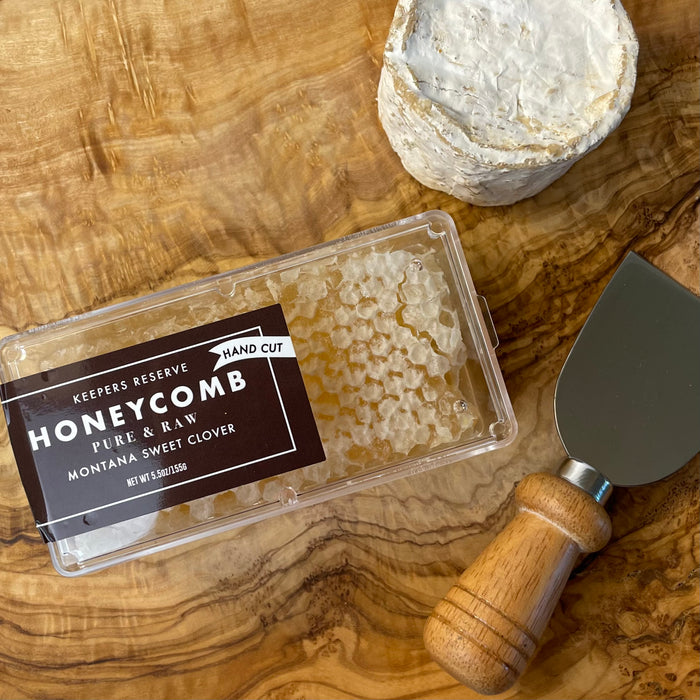 a small clear plastic box of honeycomb sits on an olivewood board with a small round of truffled brie and a cheese knife nearby.