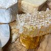 Close up of honeycomb with honey oozing out of the corner. various cheeses are shown out of focus in the background.