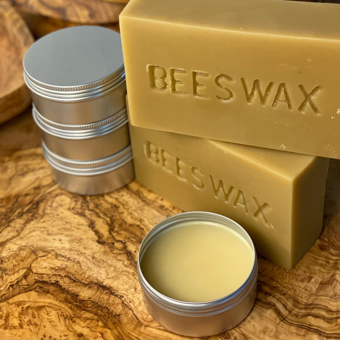 Two blocks of beeswax with the word BEESWAX are stacked on an olivewood board with four tins of wood butter; the one in front is open.