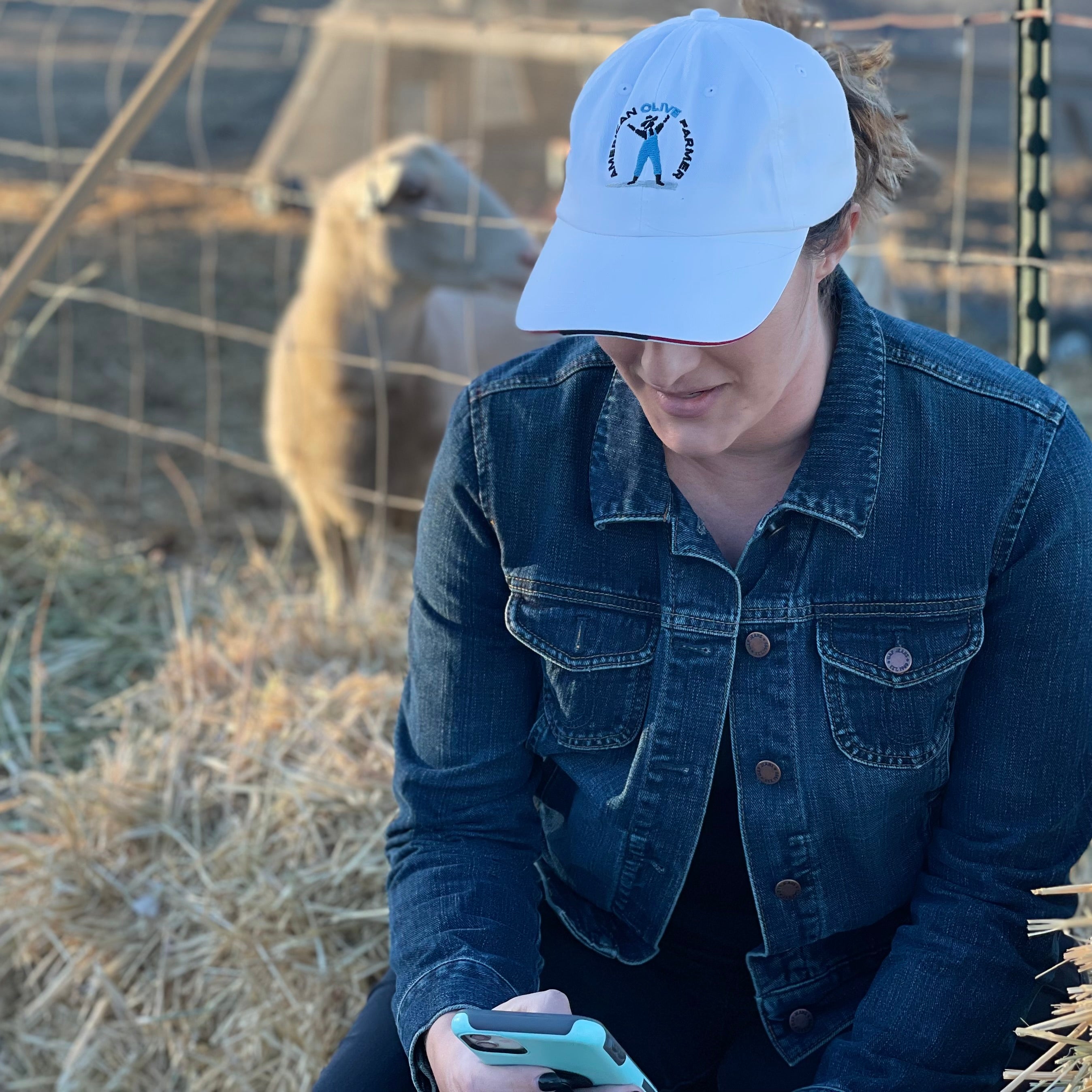 Young woman checks her phone while doing farm chores. She's wearing the white American Olive Farmer hat with red and blue accents.