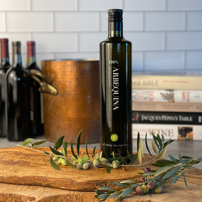 Tall bottle of Arbequina EVOO sits on olivewood with fresh arbequina olives. Copper pot and cookbooks in the background.