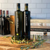 Our two heaviest and tallest bottles, 750 ml each of Arbequina EVOO and Traditional Balsamic Vinegar, stand side by side on an olivewood cutting board surrounded by arbequina olive branches full of fruit. Classic cookbooks, a hand hammered copper stock pot, and three bottles of red wine are all out of focus in the distance.