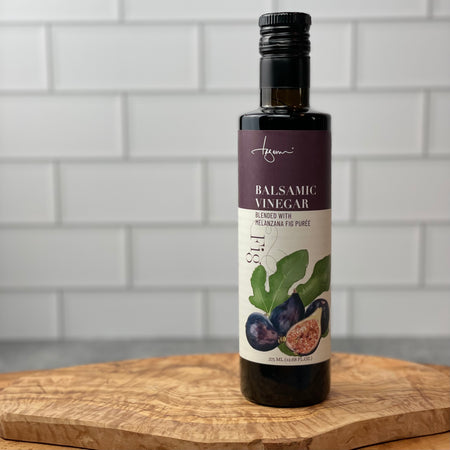 Single bottle of Melanzana Fig Balsamic Vinegar sits on an olivewood board in a white tile kitchen.