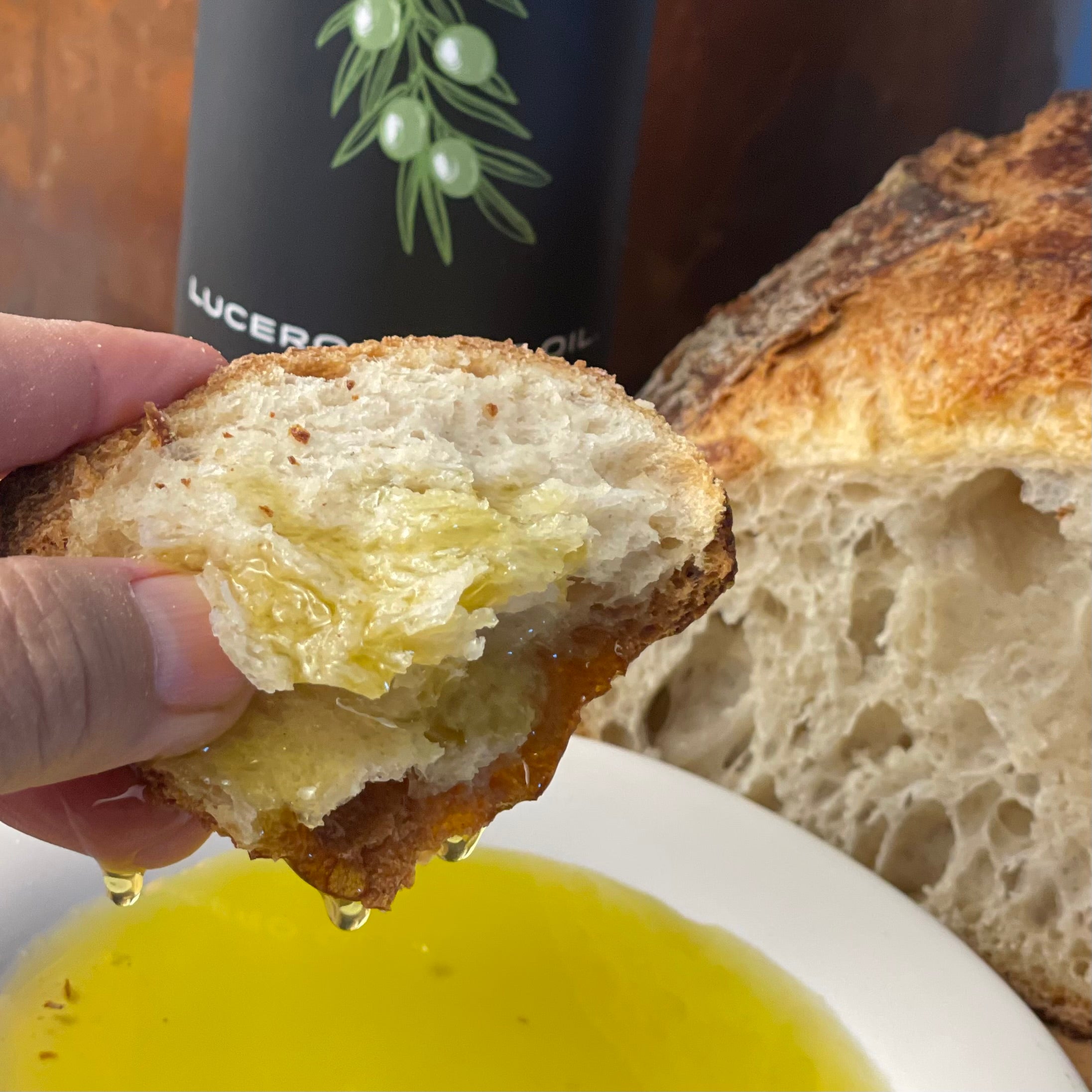 A copious amount of oil drips off a piece of chewy artisan bread shown in extreme close up. Part of the Olio Novello label as well as the hard crusty loaf is visible in the background.