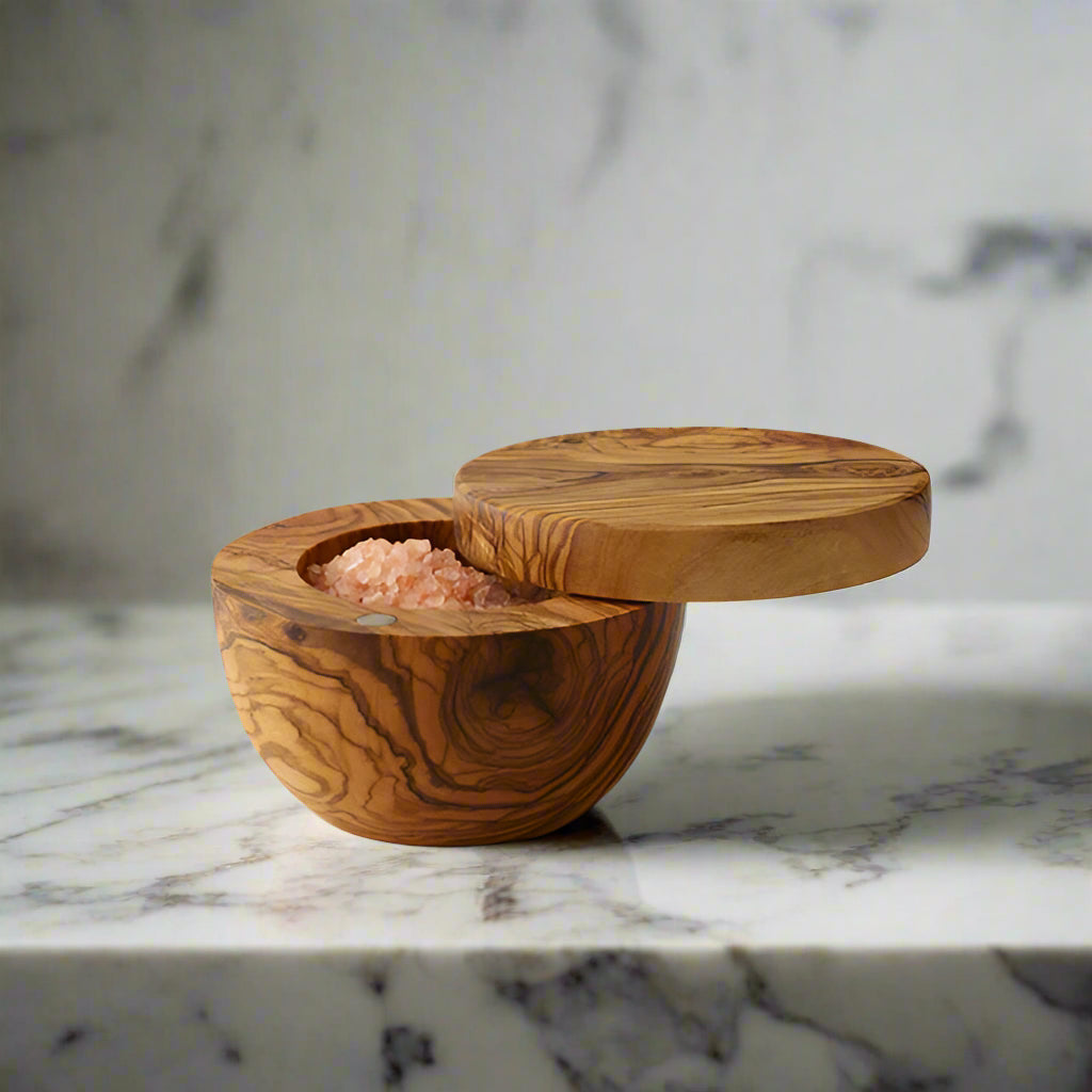 A round olive wood salt box is shown with the hinged lid swung open to reveal coarse grained pink Himalayan salt inside. The salt box sits on a dramatic white marble counter.