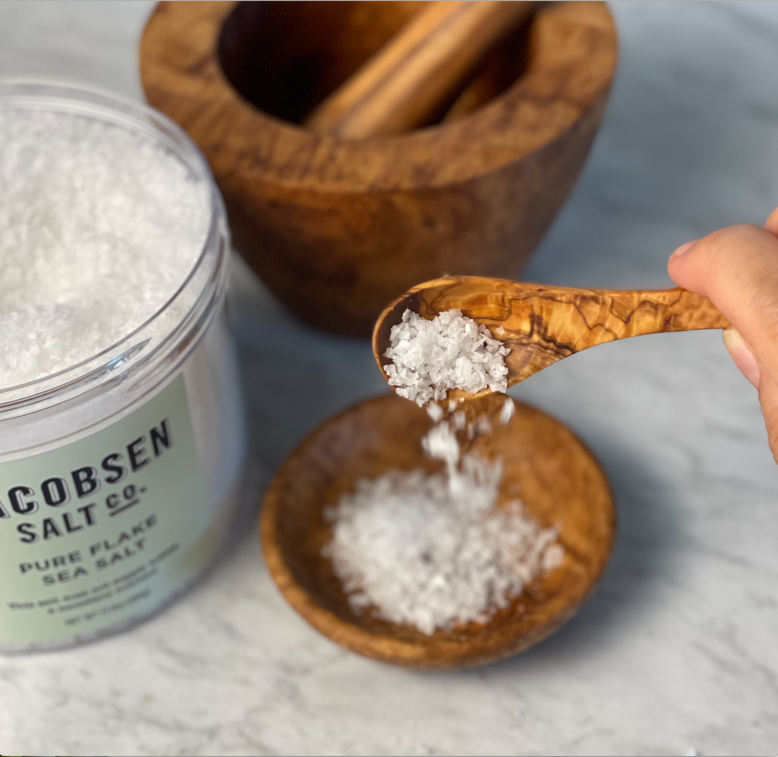 A large open jar of salt flakes sits next to an olive wood mortar and pestle. A scoop of salt falls from a small olivewood spoon into an olivewood dipping bowl.